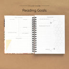 Open spiral-bound hardcover reading journal notebook showing reading goals and ratings guide on a brown background