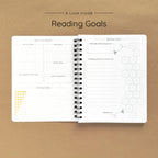Open spiral-bound softcover reading journal notebook showing reading goals and ratings guide on a brown background