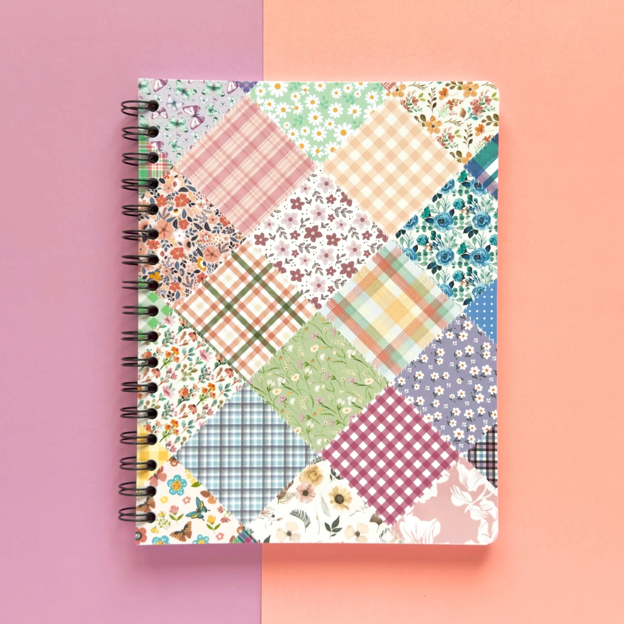 Spiral-bound softcover lined notebook with floral patchwork cover design on a lavender and peach background, featuring black wire binding