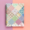 Spiral-bound hardcover lined notebook with floral patchwork cover design on a lavender and peach background