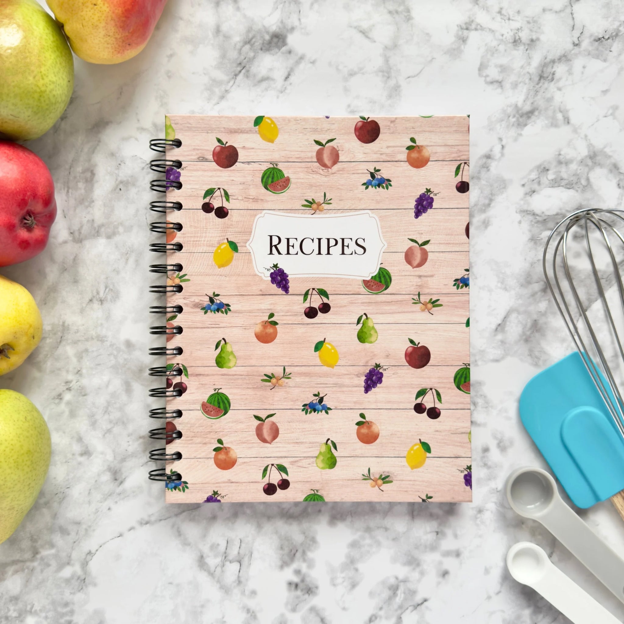 Spiral-bound hardcover recipe book notebook with fruit pattern cover design on a marble surface with kitchen utensils and pears