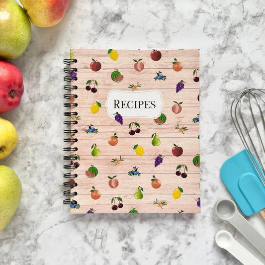 Spiral-bound hardcover recipe book notebook with fruit pattern cover design on a marble surface with kitchen utensils and pears